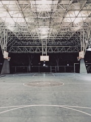 An empty indoor basketball court with a high, metal mesh ceiling and soft lighting. The court floor is painted with standard basketball markings, and there are multiple basketball hoops with backboards positioned around the court.