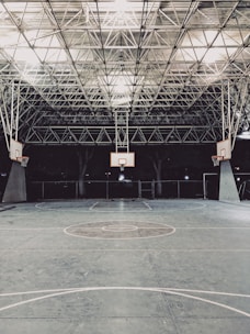 An empty indoor basketball court with a high, metal mesh ceiling and soft lighting. The court floor is painted with standard basketball markings, and there are multiple basketball hoops with backboards positioned around the court.
