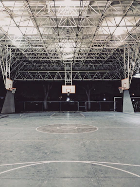 An empty indoor basketball court with a high, metal mesh ceiling and soft lighting. The court floor is painted with standard basketball markings, and there are multiple basketball hoops with backboards positioned around the court.