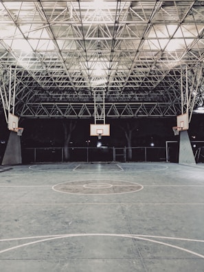 An empty indoor basketball court with a high, metal mesh ceiling and soft lighting. The court floor is painted with standard basketball markings, and there are multiple basketball hoops with backboards positioned around the court.