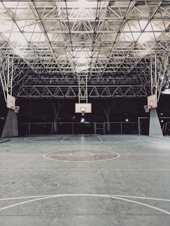 An empty indoor basketball court with a high, metal mesh ceiling and soft lighting. The court floor is painted with standard basketball markings, and there are multiple basketball hoops with backboards positioned around the court.