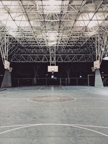 An empty indoor basketball court with a high, metal mesh ceiling and soft lighting. The court floor is painted with standard basketball markings, and there are multiple basketball hoops with backboards positioned around the court.