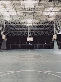 An empty indoor basketball court with a high, metal mesh ceiling and soft lighting. The court floor is painted with standard basketball markings, and there are multiple basketball hoops with backboards positioned around the court.