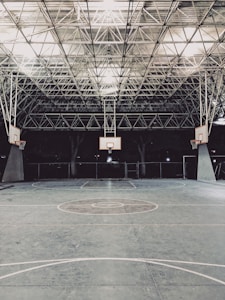 An empty indoor basketball court with a high, metal mesh ceiling and soft lighting. The court floor is painted with standard basketball markings, and there are multiple basketball hoops with backboards positioned around the court.