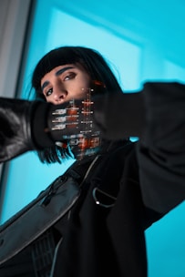 A person with dark hair and a septum piercing is wearing a futuristic outfit with a long sleeve, black jacket, and a crossbody bag. The individual has a stern look, with digital graphics and code reflections visible on their face and clothes. The background is a bright, vivid blue, adding a sci-fi ambiance.