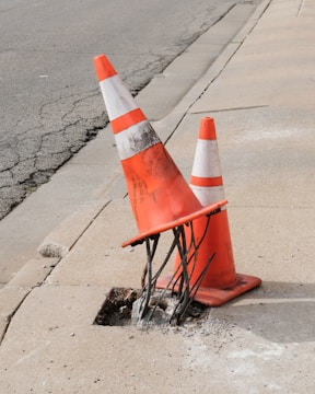 Two orange traffic cones are positioned on a concrete sidewalk next to a damaged section containing exposed rebar and a small hole. The cones have visible dirt marks on them and one is leaning over, partially submerged into the hole.