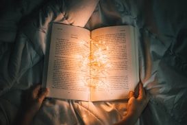 An open book is resting on a soft, wrinkled bed cover, illuminated by a string of warm, glowing fairy lights. Two hands gently hold the book open as the lights create a cozy and intimate ambiance.
