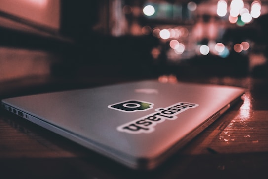 A closed laptop with a sticker on the lid, resting on a wooden surface in a dimly lit environment. The background features soft, out-of-focus lights creating a warm ambiance.