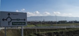 A rural road scene with a signpost indicating directions towards Pásztó and Salgótarján. The background features a landscape with open fields, suburban houses, and a church steeple, all set against a backdrop of a mountain range. The sky is clear with scattered clouds.