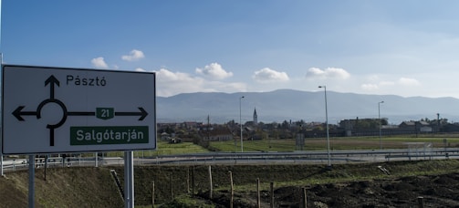 A rural road scene with a signpost indicating directions towards Pásztó and Salgótarján. The background features a landscape with open fields, suburban houses, and a church steeple, all set against a backdrop of a mountain range. The sky is clear with scattered clouds.