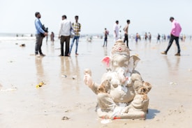 A clay statue of Lord Ganesha is partially submerged on a beach, surrounded by damp sand and some small flowers. Several people are visible in the background, some standing and others walking along the shoreline. The scene suggests a ritual or celebration taking place.