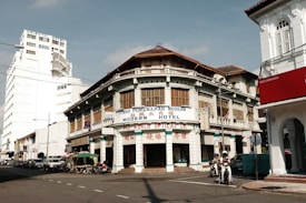 A street scene features an old-style building with signs for a modern hotel and an optical company. The architecture includes brown wood and pale green accents. Vehicles and a few people are visible on the street, including scooters. A tall white building is in the background with 'Oriental' written on it.