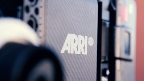 Close-up view of a professional film camera with the brand 'ARRI' prominently displayed in white on a textured black surface. The camera's components and details are visible in the background.