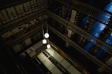 A dimly lit, multi-level library or archive featuring metal railings, shelves densely packed with books or documents, and a few hanging spherical light fixtures. The perspective offers a top-down view, highlighting the geometric patterns created by the mirrored rows of shelving and the industrial aesthetic.