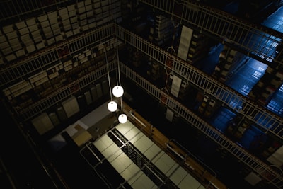 A dimly lit, multi-level library or archive featuring metal railings, shelves densely packed with books or documents, and a few hanging spherical light fixtures. The perspective offers a top-down view, highlighting the geometric patterns created by the mirrored rows of shelving and the industrial aesthetic.