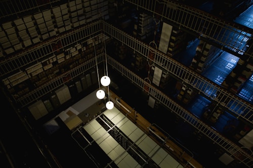 A dimly lit, multi-level library or archive featuring metal railings, shelves densely packed with books or documents, and a few hanging spherical light fixtures. The perspective offers a top-down view, highlighting the geometric patterns created by the mirrored rows of shelving and the industrial aesthetic.