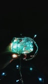 A hand holding a glass container filled with glowing string lights. The lights are emitting a soft, bluish-green glow. Additional lights are visible in the background against a dark backdrop.
