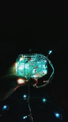 A hand holding a glass container filled with glowing string lights. The lights are emitting a soft, bluish-green glow. Additional lights are visible in the background against a dark backdrop.