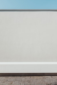A minimalist scene featuring a flat, light-colored wall with a clean, horizontal line running near its base. The foreground displays a paved surface with interlocking bricks. Above the wall, a clear blue sky completes the simple composition.