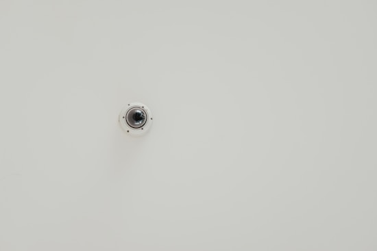A wall-mounted security camera is centered in the image against a plain, light-colored background. The camera is small, dome-shaped, and slightly reflective.