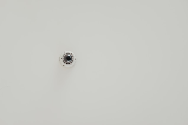 A wall-mounted security camera is centered in the image against a plain, light-colored background. The camera is small, dome-shaped, and slightly reflective.