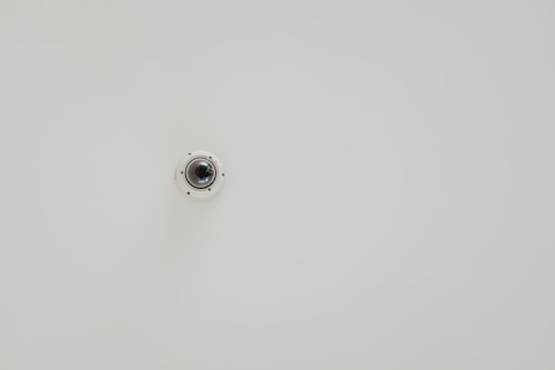 A wall-mounted security camera is centered in the image against a plain, light-colored background. The camera is small, dome-shaped, and slightly reflective.