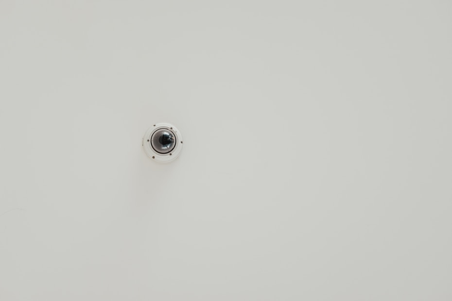 A wall-mounted security camera is centered in the image against a plain, light-colored background. The camera is small, dome-shaped, and slightly reflective.