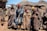 A group of six people stand together wearing traditional, colorful blankets and head coverings in front of a rural setting with thatch-roofed huts made of stone. There is a black and brown dog to the left, and two white vehicles partially visible in the background. The environment appears mountainous and sunny with a clear blue sky.
