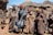 A group of six people stand together wearing traditional, colorful blankets and head coverings in front of a rural setting with thatch-roofed huts made of stone. There is a black and brown dog to the left, and two white vehicles partially visible in the background. The environment appears mountainous and sunny with a clear blue sky.