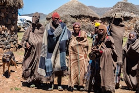 A group of six people stand together wearing traditional, colorful blankets and head coverings in front of a rural setting with thatch-roofed huts made of stone. There is a black and brown dog to the left, and two white vehicles partially visible in the background. The environment appears mountainous and sunny with a clear blue sky.
