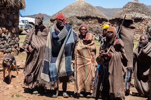 A group of six people stand together wearing traditional, colorful blankets and head coverings in front of a rural setting with thatch-roofed huts made of stone. There is a black and brown dog to the left, and two white vehicles partially visible in the background. The environment appears mountainous and sunny with a clear blue sky.