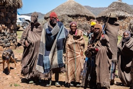 A group of six people stand together wearing traditional, colorful blankets and head coverings in front of a rural setting with thatch-roofed huts made of stone. There is a black and brown dog to the left, and two white vehicles partially visible in the background. The environment appears mountainous and sunny with a clear blue sky.