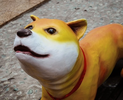 A colorful dog sculpture with a smooth texture is positioned on a pebble-like walkway. The dog has a white face with a gradual transition to yellow and orange towards its back. It sports a red collar and has a cute, wide-eyed expression.