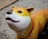 A colorful dog sculpture with a smooth texture is positioned on a pebble-like walkway. The dog has a white face with a gradual transition to yellow and orange towards its back. It sports a red collar and has a cute, wide-eyed expression.