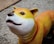 A colorful dog sculpture with a smooth texture is positioned on a pebble-like walkway. The dog has a white face with a gradual transition to yellow and orange towards its back. It sports a red collar and has a cute, wide-eyed expression.