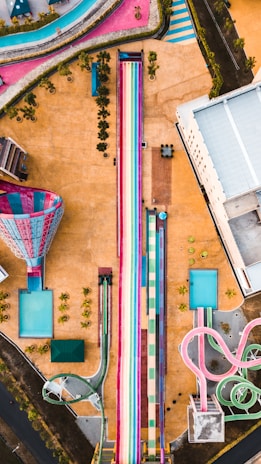An aerial view of a colorful water park featuring large water slides, including a prominent rainbow-colored slide in the center. Surrounding the slides are various pools and pathways, with some greenery and a few structures scattered throughout. The landscape is vibrant, with an array of colors giving a lively and playful feel.