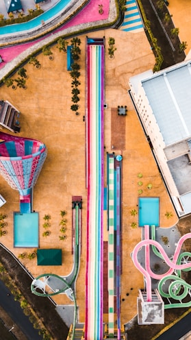 An aerial view of a colorful water park featuring large water slides, including a prominent rainbow-colored slide in the center. Surrounding the slides are various pools and pathways, with some greenery and a few structures scattered throughout. The landscape is vibrant, with an array of colors giving a lively and playful feel.