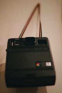A vintage instant camera with a rainbow-colored strap hanging against a plain wall. The camera is dark in color, with visible buttons and a logo featuring multiple colors.