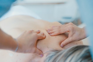 Hands performing a massage on a person's back, applying pressure to specific points. The setting is bright and calming, with soft focus creating a tranquil atmosphere.