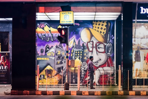 A vibrant street scene featuring a large, colorful mural painted on the storefront's shutters. The artwork includes a cartoonish character with oversized glasses and headphones, set against an urban backdrop with abstract designs and bright colors. A man stands in front of the mural, observed partially from the side. To the right, the illuminated interior of a clothing store is visible. Above, a UCO Bank sign hangs next to a traffic light displaying red.