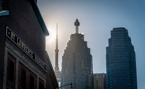 Silhouetted skyline featuring modern skyscrapers and a prominent needle-like communications tower against a bright sunlit sky. The foreground includes a brick building with a sign reading 'St. Lawrence Market.'