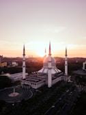 A large mosque with a blue dome and four towering minarets is set against a stunning sunset. The architecture is intricate and grand, with a spacious courtyard visible. The surrounding landscape includes lush greenery and modern buildings in the distance.