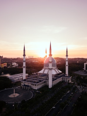 A large mosque with a blue dome and four towering minarets is set against a stunning sunset. The architecture is intricate and grand, with a spacious courtyard visible. The surrounding landscape includes lush greenery and modern buildings in the distance.