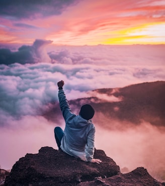 man sitting on mountain cliff facing white clouds rising one hand at golden hour