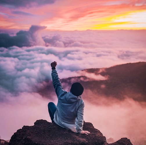 man sitting on mountain cliff facing white clouds rising one hand at golden hour