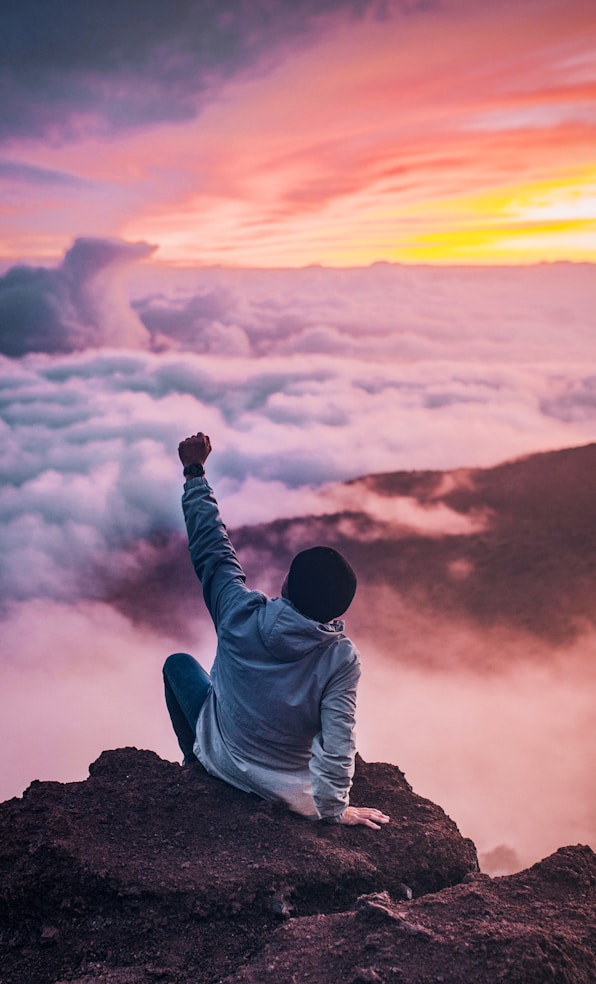 man sitting on mountain cliff facing white clouds rising one hand at golden hour