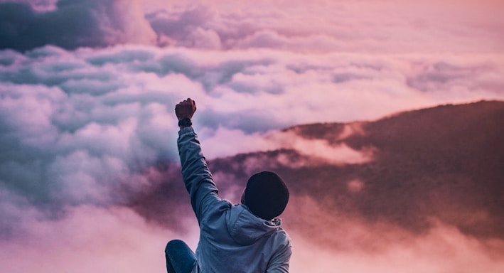 man sitting on mountain cliff facing white clouds rising one hand at golden hour