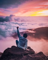 man sitting on mountain cliff facing white clouds rising one hand at golden hour
