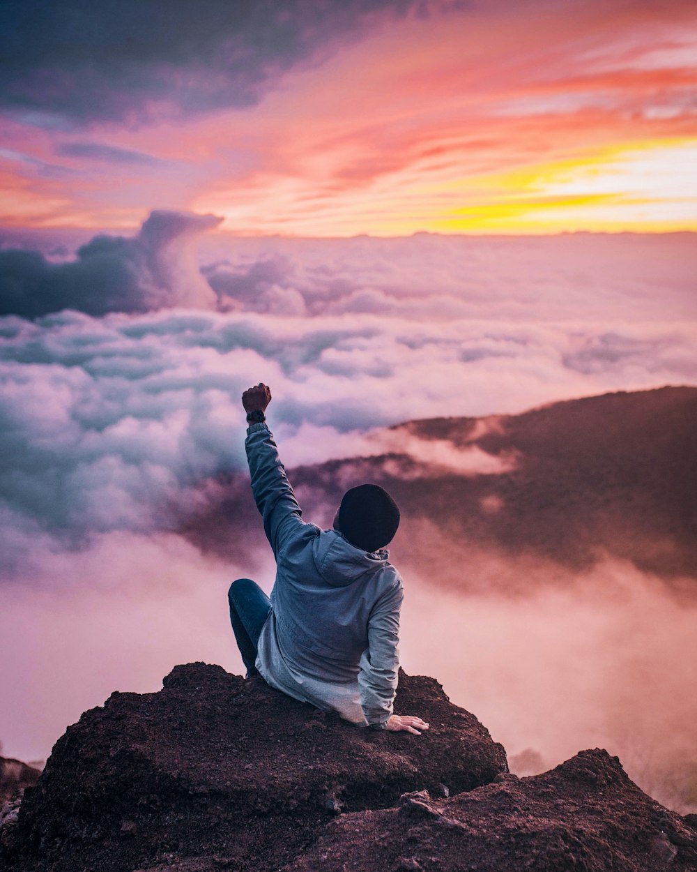 man sitting on mountain cliff facing white clouds rising one hand at golden hour