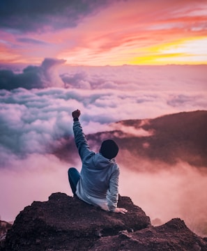 man sitting on mountain cliff facing white clouds rising one hand at golden hour